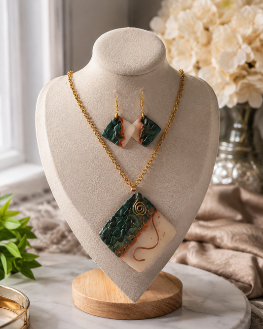 Conjunto de joyas expuesto en un busto color crema, compuesto por un collar de cadena dorada con un colgante rectangular y pendientes colgantes a juego. Las joyas presentan secciones de piedra jaspeada verde oscuro, secciones de piedra rosa melocotón y secciones de piedra color crema con un pequeño detalle en espiral dorada - D'Elle Gifts