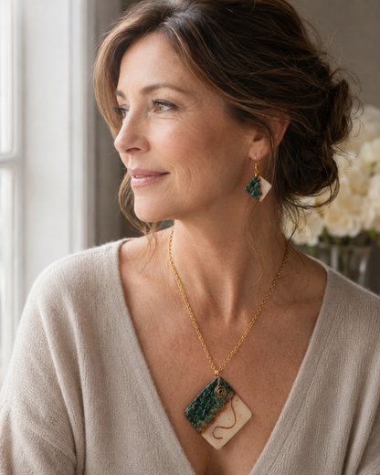 Una mujer luce un collar de cadena de oro con un colgante rectangular con un estampado jaspeado en verde y crema, y ​​un pendiente colgante a juego, en un ambiente interior con iluminación tenue - D'Elle Gifts