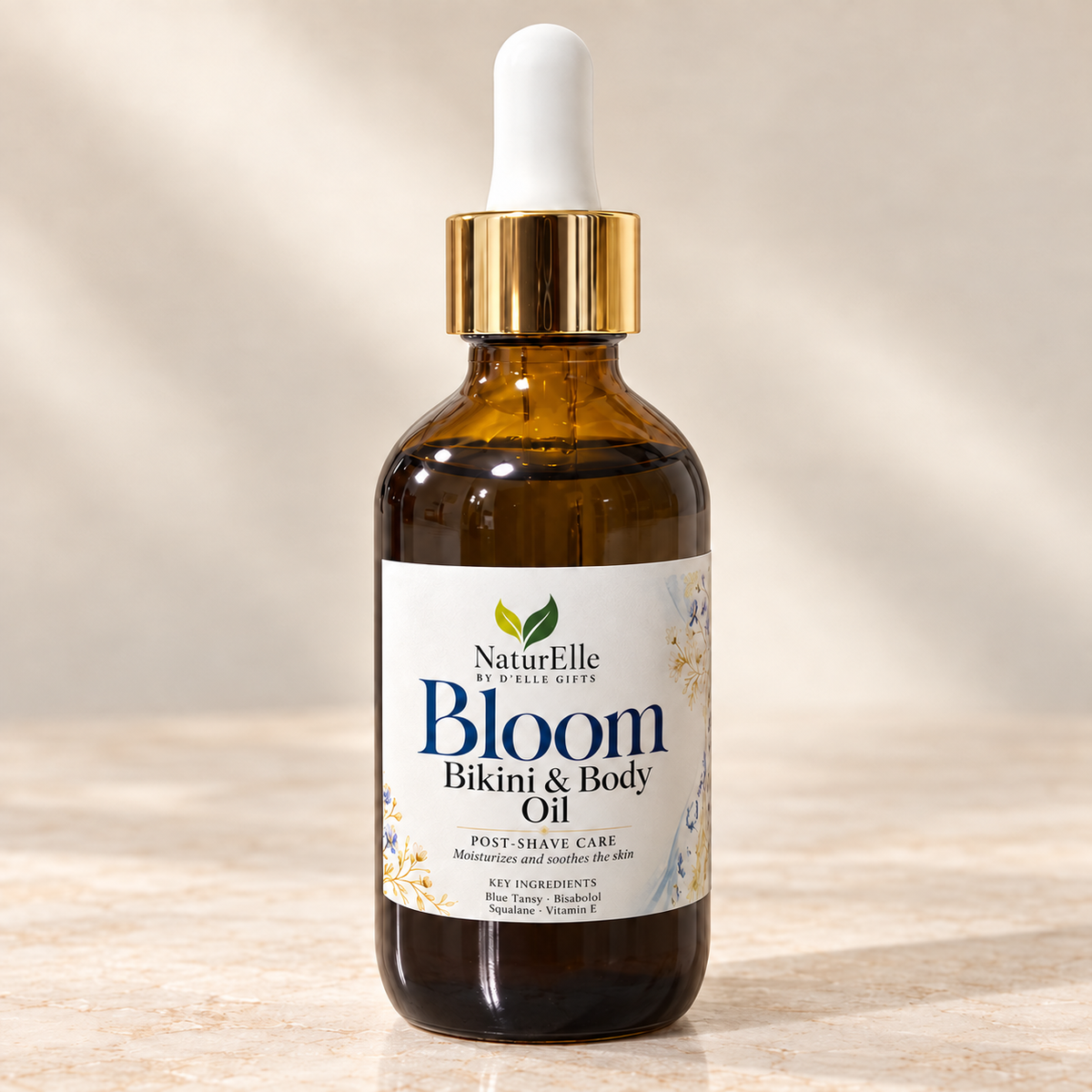 A bottle of NaturElle by Ellie Gifts Bloom Bikini & Body Oil post-shave care, in an amber glass bottle with a gold collar and white dropper cap, set on a light marble surface with a warm beige background.