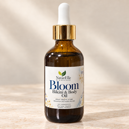 A bottle of NaturElle by Ellie Gifts Bloom Bikini & Body Oil post-shave care, in an amber glass bottle with a gold collar and white dropper cap, set on a light marble surface with a warm beige background.
