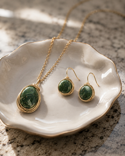 Conjunto de joyas de oro con collar colgante y pendientes a juego, con cabujones ovalados de piedra verde engastados en oro, presentados sobre un pequeño plato de cerámica blanca sobre una superficie de piedra moteada - D'Elle Gifts