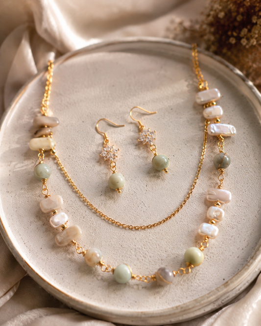 Conjunto de joyas en tono dorado compuesto por un collar de cuentas y pendientes colgantes a juego, presentado sobre un plato redondo de cerámica con tela beige suave y flores secas de fondo - D'Elle Gifts