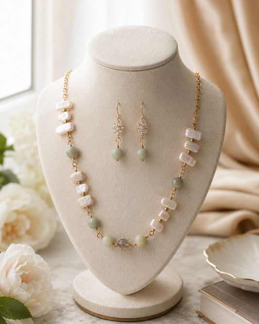 Conjunto de joyas en tono dorado, presentado sobre un busto color crema, compuesto por un collar y pendientes colgantes a juego. El collar y los pendientes están decorados con cuentas de concha blancas de estilo barroco, cuentas redondas verde claro y detalles de cristal transparente - D'Elle Gifts