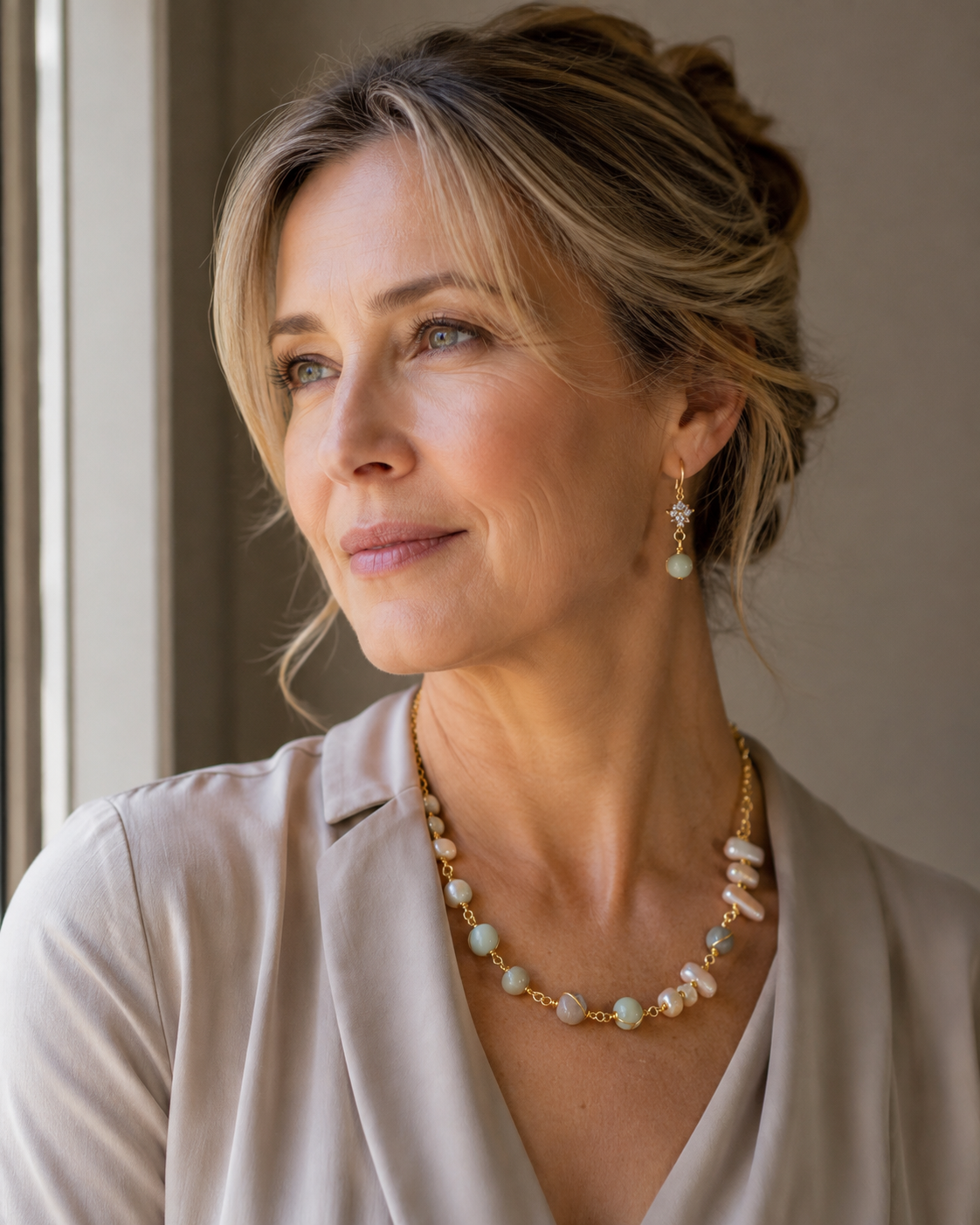 Una mujer rubia con el pelo recogido, luciendo un collar de oro y perlas y pendientes colgantes a juego, vestida con una blusa beige claro, fue capturada en un retrato cálido y desenfocado - D'Elle Gifts