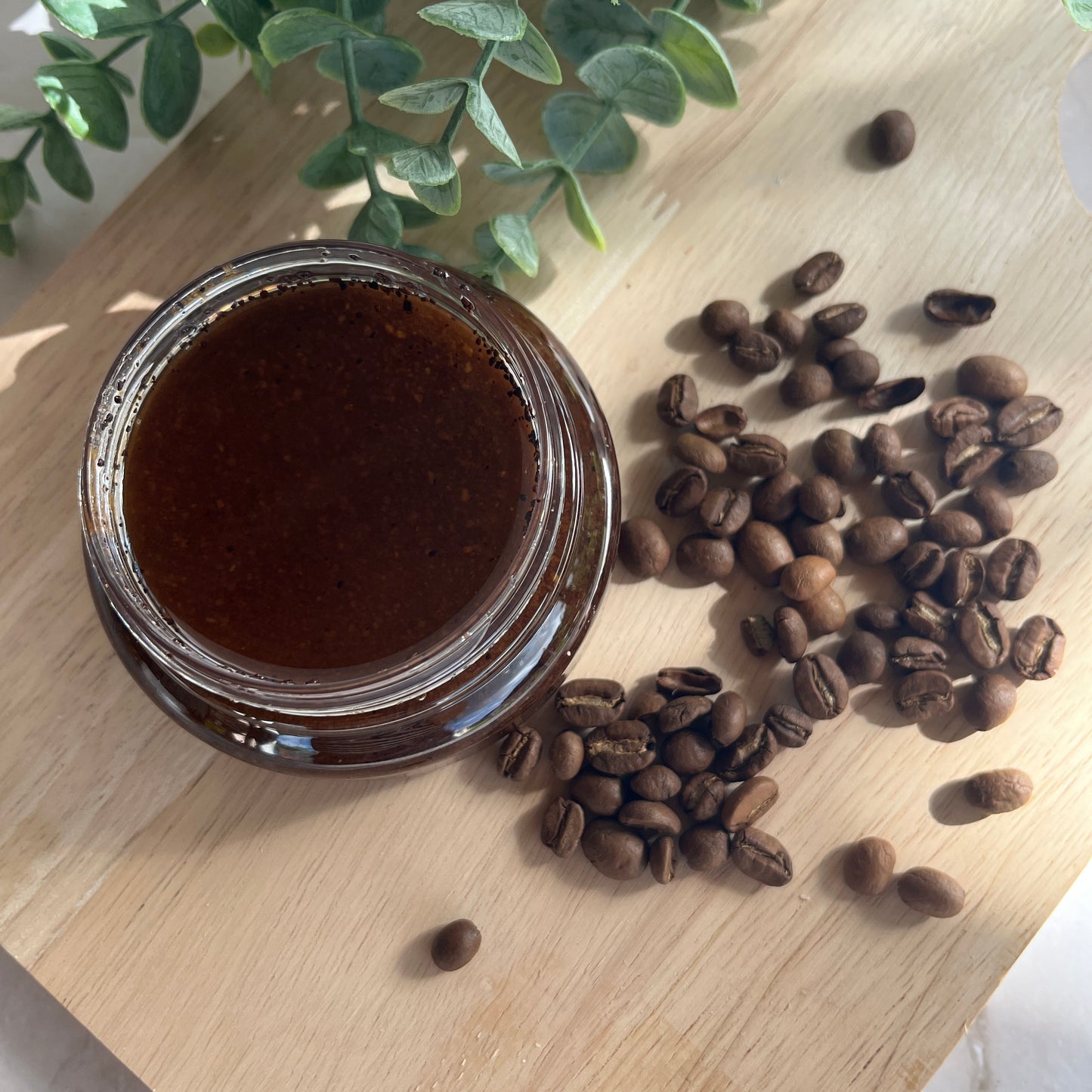 Jar of dark liquid next to coffee beans on a wooden surface with a plant in the background