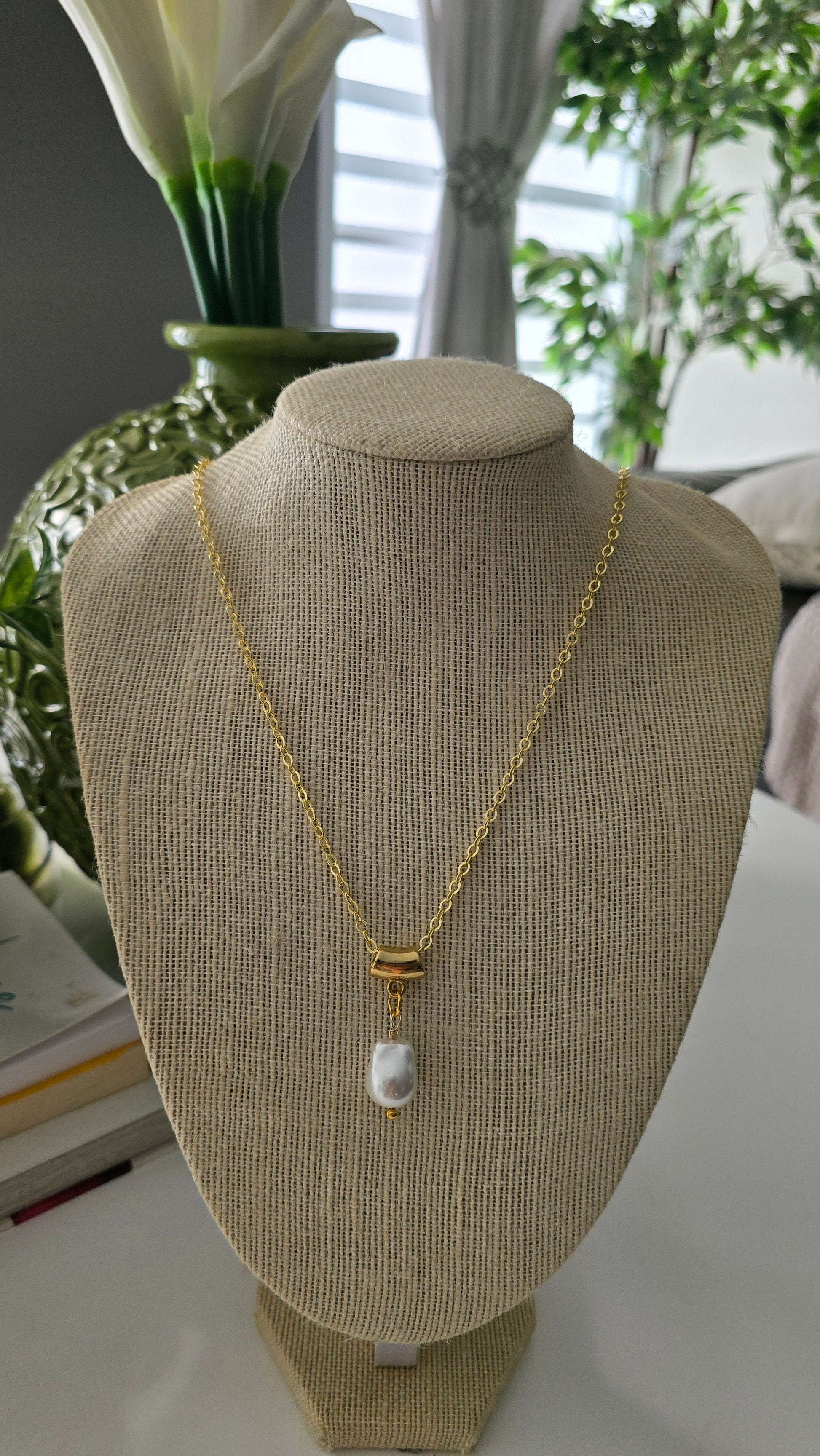 Cabeza de maniquí con superficie texturizada beige y cadena dorada con una pequeña perla sobre un fondo interior difuminado.