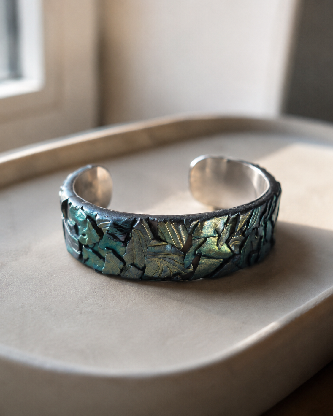 Una pulsera de plata con un diseño texturizado de hojas superpuestas en tonos iridiscentes verdes y azules, sobre una bandeja beige - D'Elle Gifts