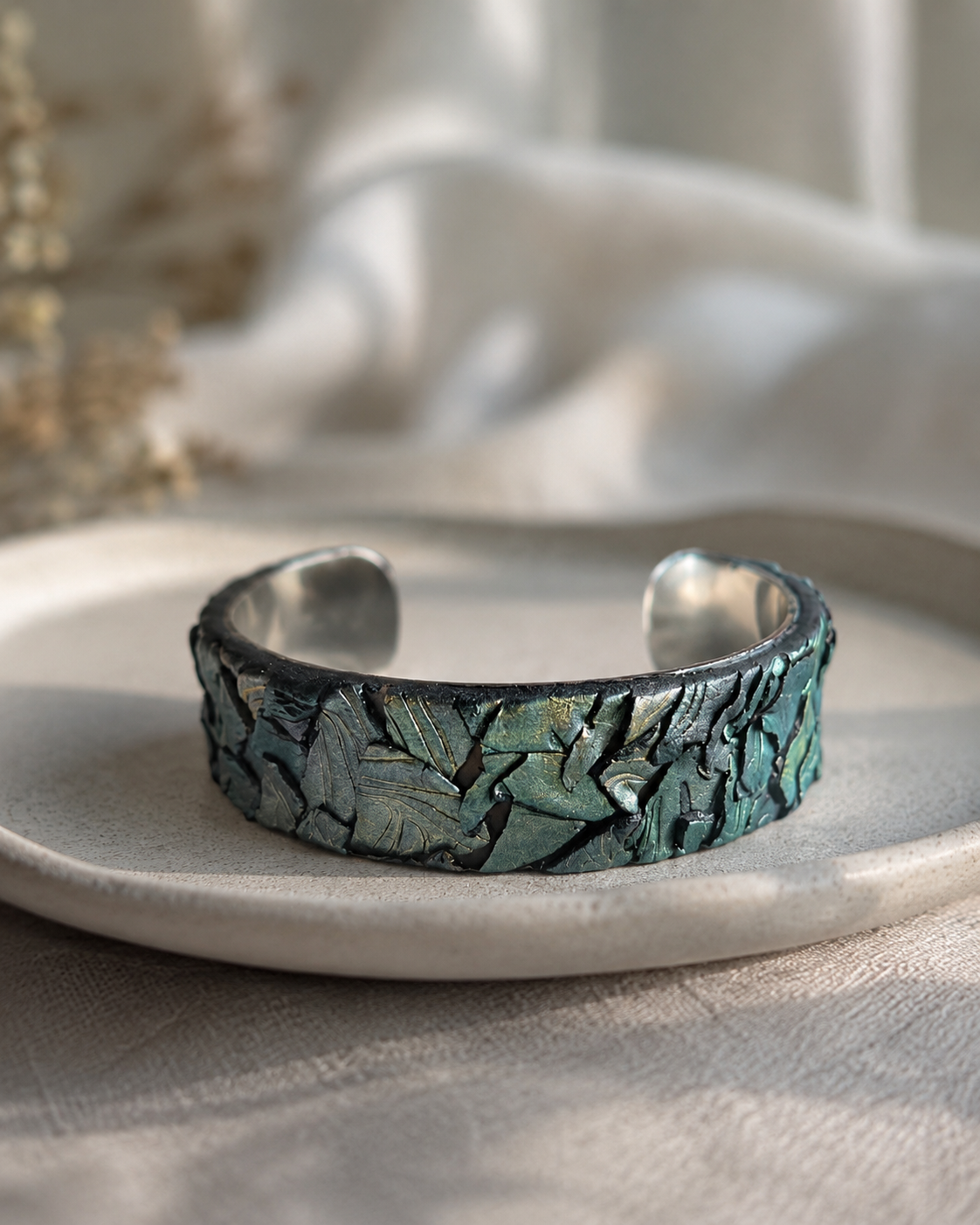 Pulsera abierta plateada con una banda texturizada de hojas metálicas en tonos verde azulado, verde y bronce, sobre un plato de cerámica gris claro - D'Elle Gifts