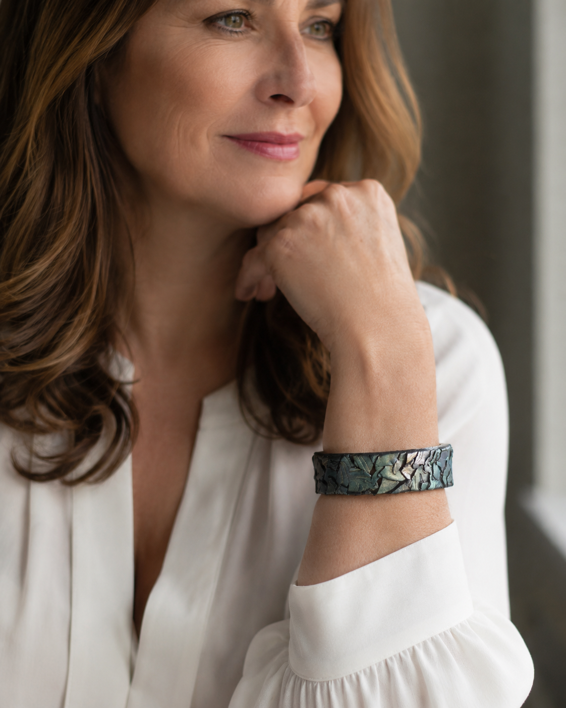 Una mujer con blusa blanca y una pulsera ancha con estampado de hojas azul oscuro y gris, apoya la barbilla en la mano en un ambiente interior con iluminación tenue - D'Elle Gifts