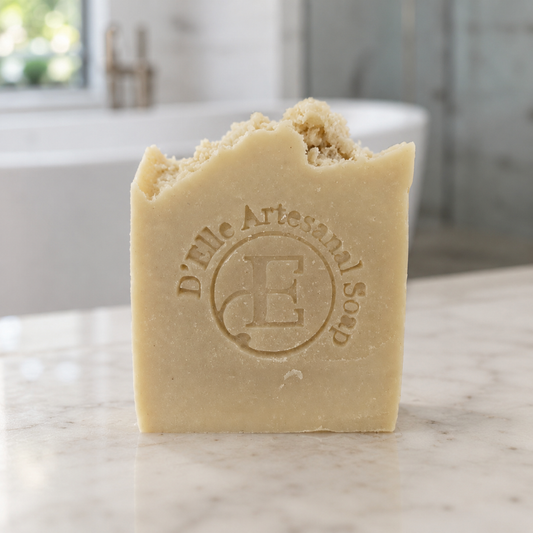 Una barra rectangular de jabón artesanal D'Elle con el borde superior irregular y fragmentado, apoyada sobre una encimera de mármol blanco y con un fondo de baño desenfocado — NaturElle Puerto Rico