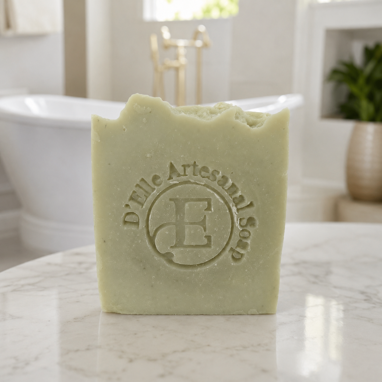 Una barra rectangular de jabón artesanal verde pálido se yergue sobre una superficie de mármol blanco, con una escena de baño borrosa al fondo. El jabón lleva grabado el texto "D'Elle Artesanal Soap" y una gran letra "E" — NaturElle Puerto Rico