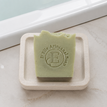Una barra de jabón rectangular de color verde pálido con un borde superior rústico y roto, sobre una pequeña jabonera blanquecina. El jabón lleva grabado el nombre de la marca "D'Elle Artesanal Soap" y un monograma — NaturElle Puerto Rico