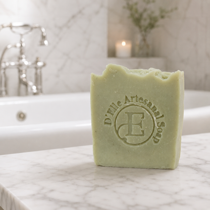 Una barra rectangular de jabón verde pálido sobre una superficie de mármol blanco, con una escena de baño suavemente difuminada al fondo. El jabón lleva grabado el nombre de la marca «D'Elle Artesanal Soap» y un monograma — NaturElle Puerto Rico