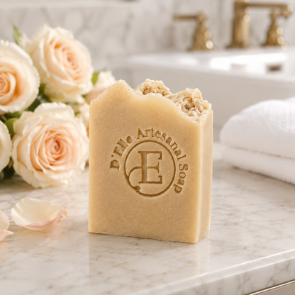 Una barra rectangular de jabón artesanal D'Elle sobre una encimera de mármol en el baño, acompañada de rosas color melocotón pálido y una toalla blanca doblada, con un lavado suavemente desenfocado al fondo — NaturElle Puerto Rico