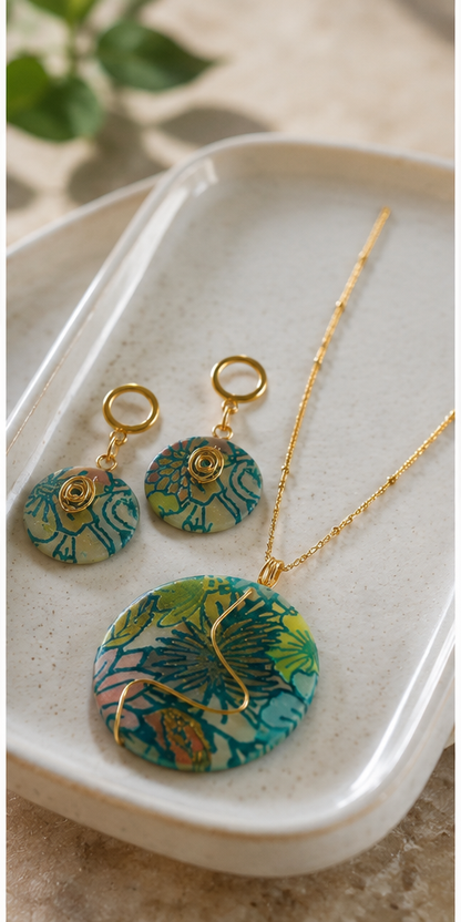 Un collar de oro y pendientes colgantes a juego con colgantes redondos estampados con motivos florales en verde, turquesa y naranja, presentados en una bandeja de cerámica beige - D'Elle Gifts