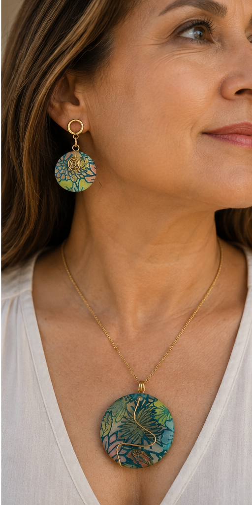 Una mujer luce un collar de oro con un colgante floral redondo y pendientes colgantes a juego, en un retrato de primer plano. La joya presenta un estampado floral tropical multicolor con tonos azules, verdes y amarillos sobre una montura dorada - D'Elle Gifts