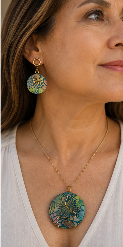 Una mujer luce un collar de oro con un colgante floral redondo y pendientes colgantes a juego, en un retrato de primer plano. La joya presenta un estampado floral tropical multicolor con tonos azules, verdes y amarillos sobre una montura dorada - D'Elle Gifts