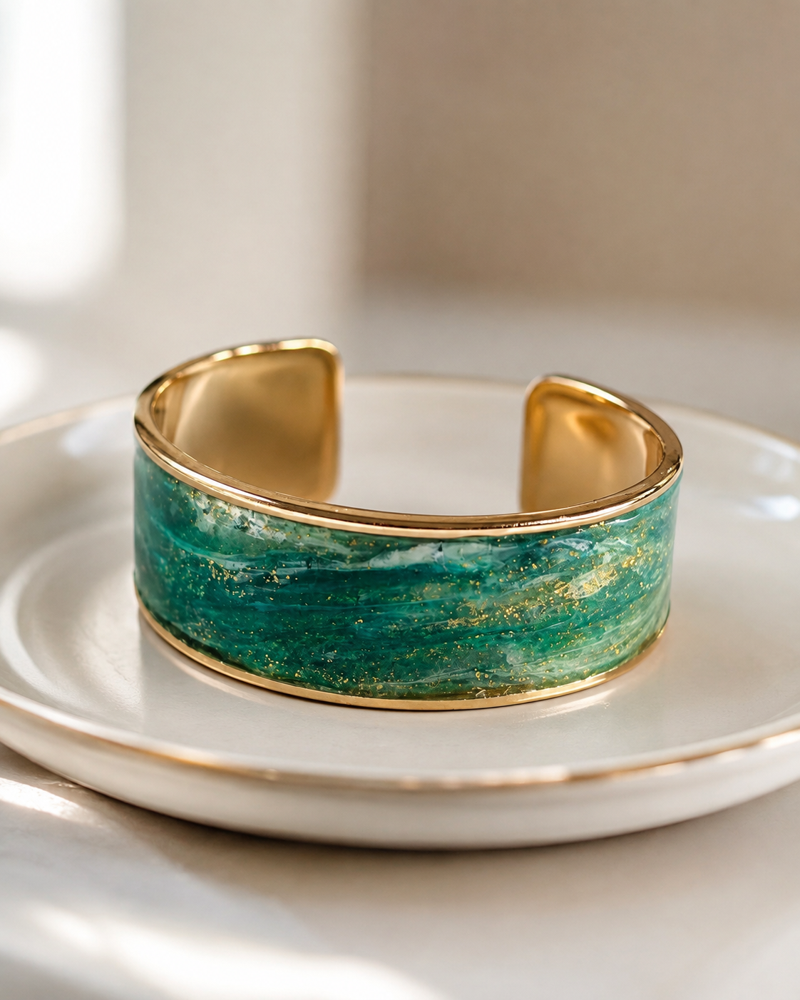 Una pulsera ancha tipo brazalete con borde de metal dorado y centro de esmalte verde texturizado, sobre una placa de cerámica blanca con ribete dorado - D'Elle Gifts