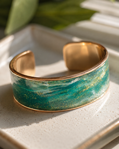 Una pulsera ancha tipo brazalete con borde de metal dorado pulido e incrustaciones de resina texturizada en tonos verde y turquesa salpicadas de purpurina dorada, sobre una bandeja de cerámica beige claro - D'Elle Gifts