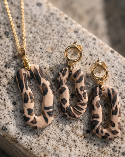 Un collar de oro y pendientes colgantes a juego con colgantes irregulares en forma de anillo con estampado animal en beige y negro, expuestos sobre una superficie de piedra moteada - D'Elle Gifts