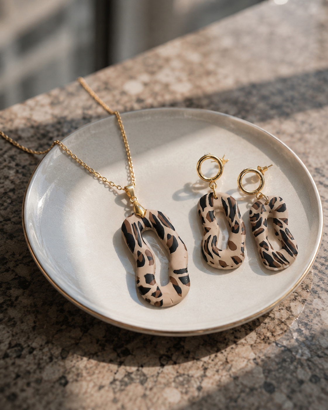 Un collar de oro y unos pendientes colgantes a juego con colgantes de estampado de leopardo en beige y negro, expuestos en un pequeño plato de cerámica blanca sobre una encimera de granito - D'Elle Gifts