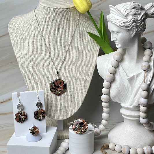 Un conjunto de joyas hechas a mano que incluye un collar, pendientes, anillo y pulsera en tonos tierra, expuestos sobre un busto de maniquí y un soporte blanco, con un plato para anillos y un objeto decorativo al fondo