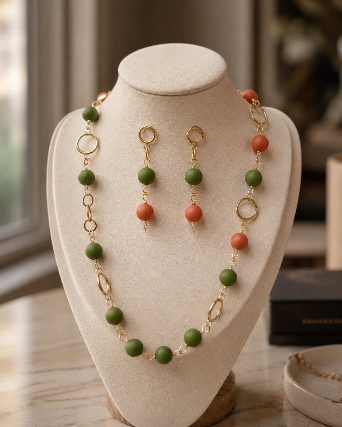 Conjunto de joyas expuesto en un busto color crema, compuesto por un collar y pendientes colgantes a juego con cuentas verdes, cuentas color coral-anaranjado y eslabones y anillos de metal dorado - D'Elle Gifts