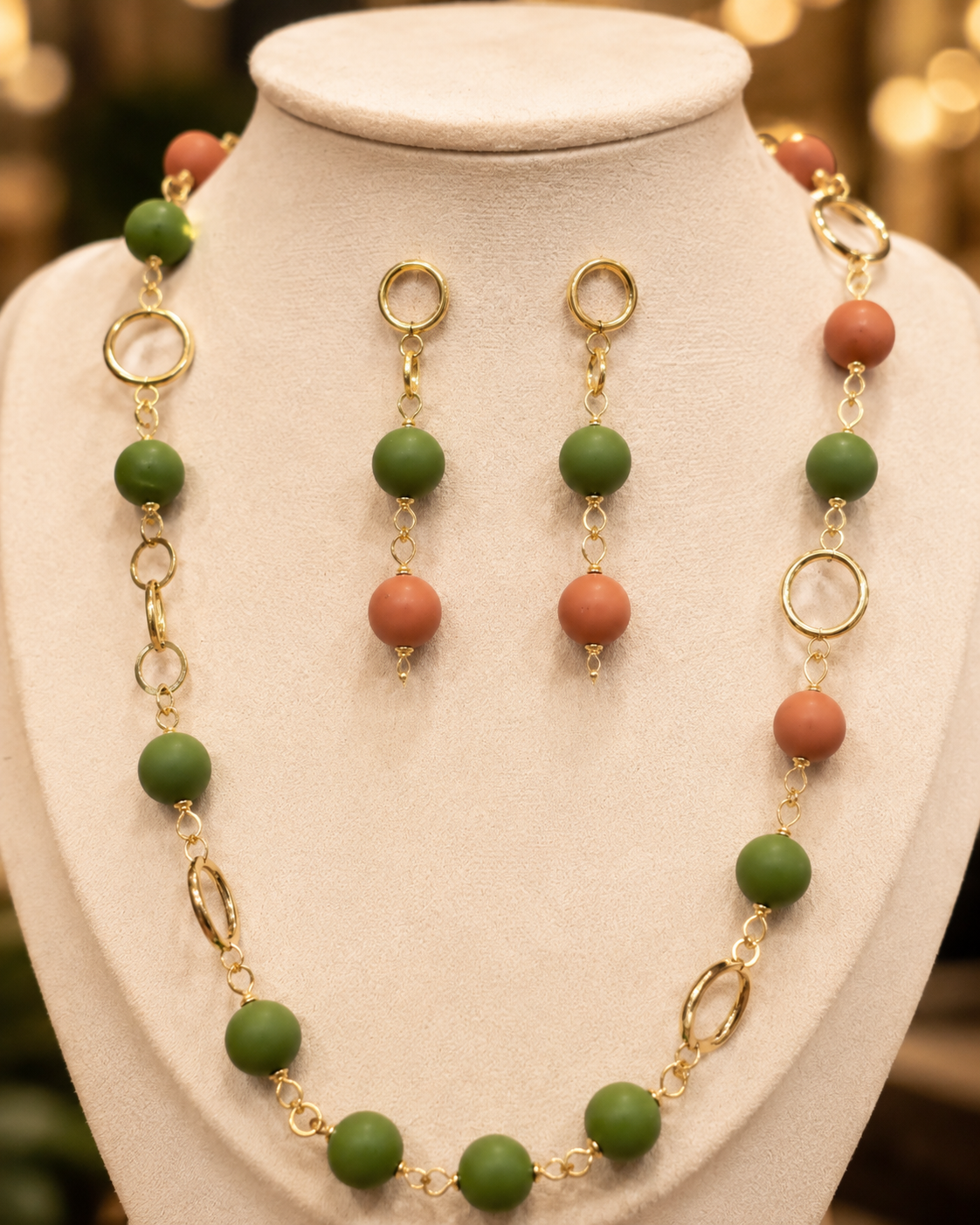 Un conjunto de joyas exhibido en un maniquí color crema, compuesto por un collar de cuentas y pendientes colgantes a juego. Las cuentas alternan entre verde mate y naranja terracota, unidas por eslabones de cadena dorados y colgantes ovalados. - D'Elle Gifts