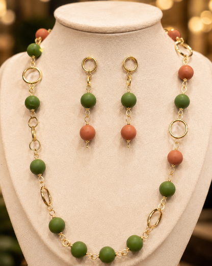 Un conjunto de joyas exhibido en un maniquí color crema, compuesto por un collar de cuentas y pendientes colgantes a juego. Las cuentas alternan entre verde mate y naranja terracota, unidas por eslabones de cadena dorados y colgantes ovalados. - D'Elle Gifts